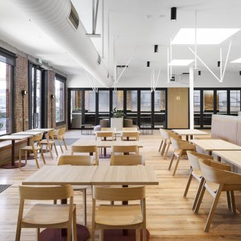 Yarra Falls, Melbourne, featuring a premium workplace café and collaboration lounge with exposed heritage brick walls, polished timber flooring, contemporary timber furniture, upholstered banquette seating, and abundant natural light, creating a welcoming environment for informal meetings, team collaboration, dining, and employee wellbeing within a character-rich commercial precinct.
