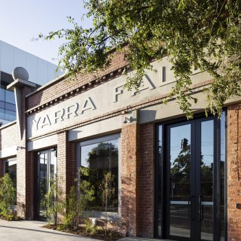 Yarra Falls, Melbourne, featuring a restored heritage industrial building with exposed red-brick masonry, original architectural detailing, contemporary glazed entrances, and prominent Yarra Falls branding. Framed by mature street trees and landscaped planting, the building reflects the successful integration of heritage character and modern workplace design within a premium commercial campus.