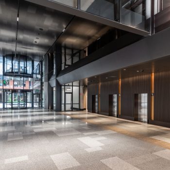 Industry Lanes, Melbourne, featuring a dramatic premium office lobby with destination-controlled lift banks, double-height ceilings, polished terrazzo flooring, exposed heritage brickwork, black steel architectural detailing, and contemporary workplace design that creates a sophisticated arrival experience within a landmark mixed-use commercial precinct.