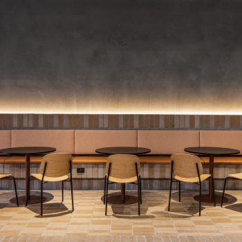 Industry Lanes, Melbourne, featuring a premium workplace collaboration lounge with integrated banquette seating, round café-style tables, timber dining chairs, exposed brickwork, textured masonry detailing, and warm ambient lighting, creating a hospitality-inspired environment that supports informal meetings, workplace connection, and tenant wellbeing within a heritage-industrial commercial precinct.