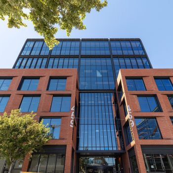 Industry Lanes, Melbourne, featuring a landmark commercial office building with a distinctive red-brick façade, expansive floor-to-ceiling glazing, recessed window openings, and mature streetscape landscaping. The contemporary workplace destination combines industrial-inspired architecture with premium office accommodation, creating a strong presence within Melbourne’s inner-city commercial precinct.